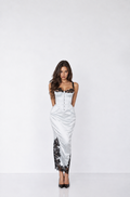 A woman modeling a white satin-like maxi dress with black lace trim at the bust and along one leg, standing against a plain light gray studio background.