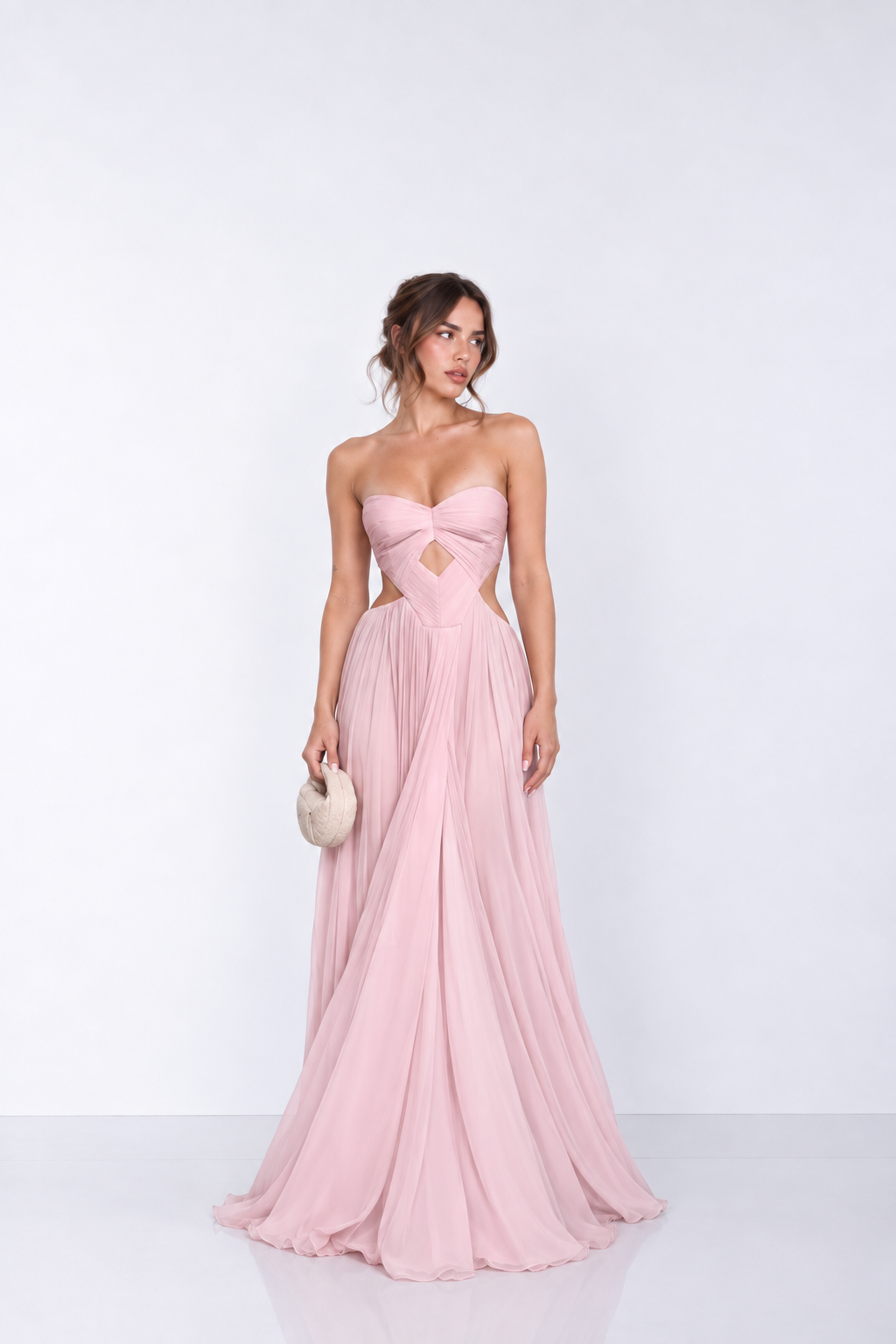 A woman modeling a strapless pink floor-length gown with a sweetheart neckline, side cutouts, and a flowing chiffon skirt, standing against a plain light gray studio background.