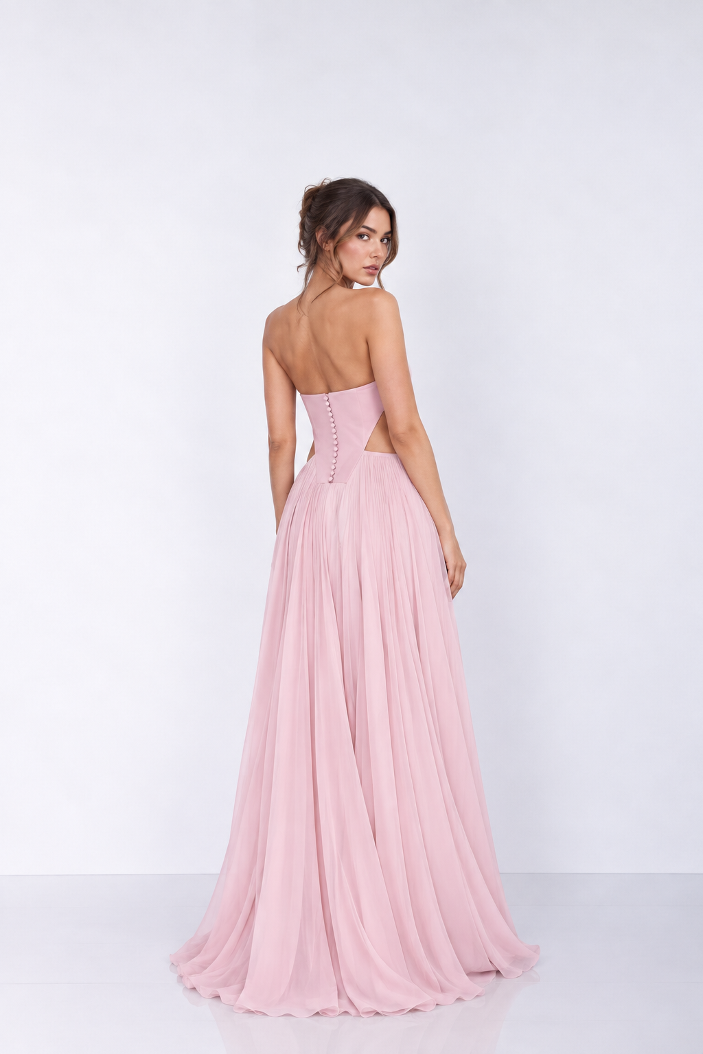 A woman modeling a strapless pink formal gown from the back, showing a structured sweetheart-style back bodice with button detailing and a flowing chiffon skirt, against a plain white studio background.u