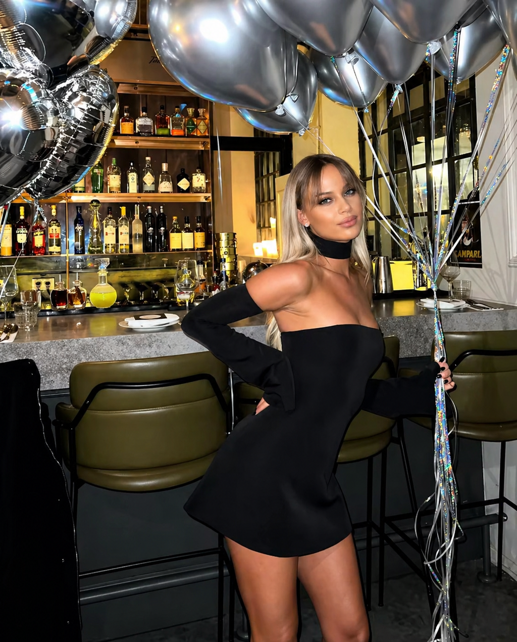 Woman in a black dress standing in a bar with balloons and bottles in the background