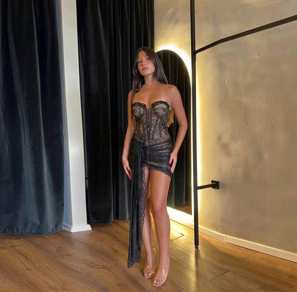 Woman in a lace outfit standing in front of a mirror in a room with wooden flooring and black curtains.