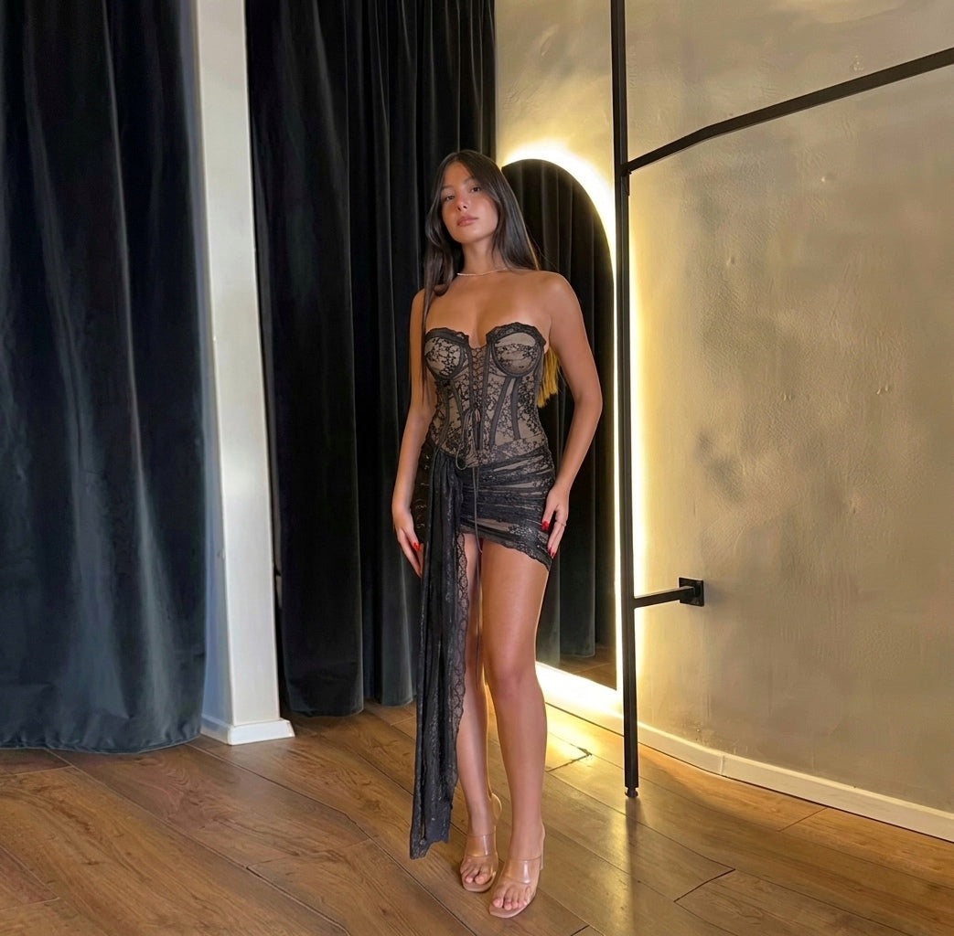 Woman in a lace outfit standing in front of a mirror in a room with wooden flooring and black curtains.