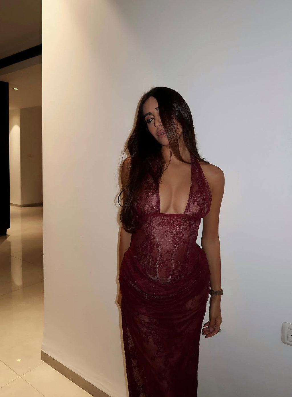 Woman in a burgundy lace dress standing against a white wall.