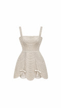 An off-white textured lace dress with a sweetheart neckline, thin shoulder straps, a fitted bodice, and a flared skirt with scalloped edges and small bow details at the hips, shown against a plain white background.