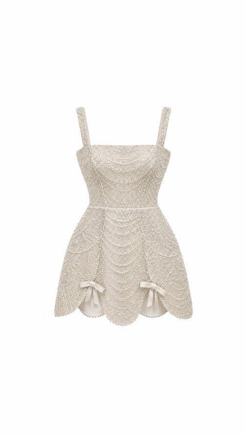 An off-white textured lace dress with a sweetheart neckline, thin shoulder straps, a fitted bodice, and a flared skirt with scalloped edges and small bow details at the hips, shown against a plain white background.