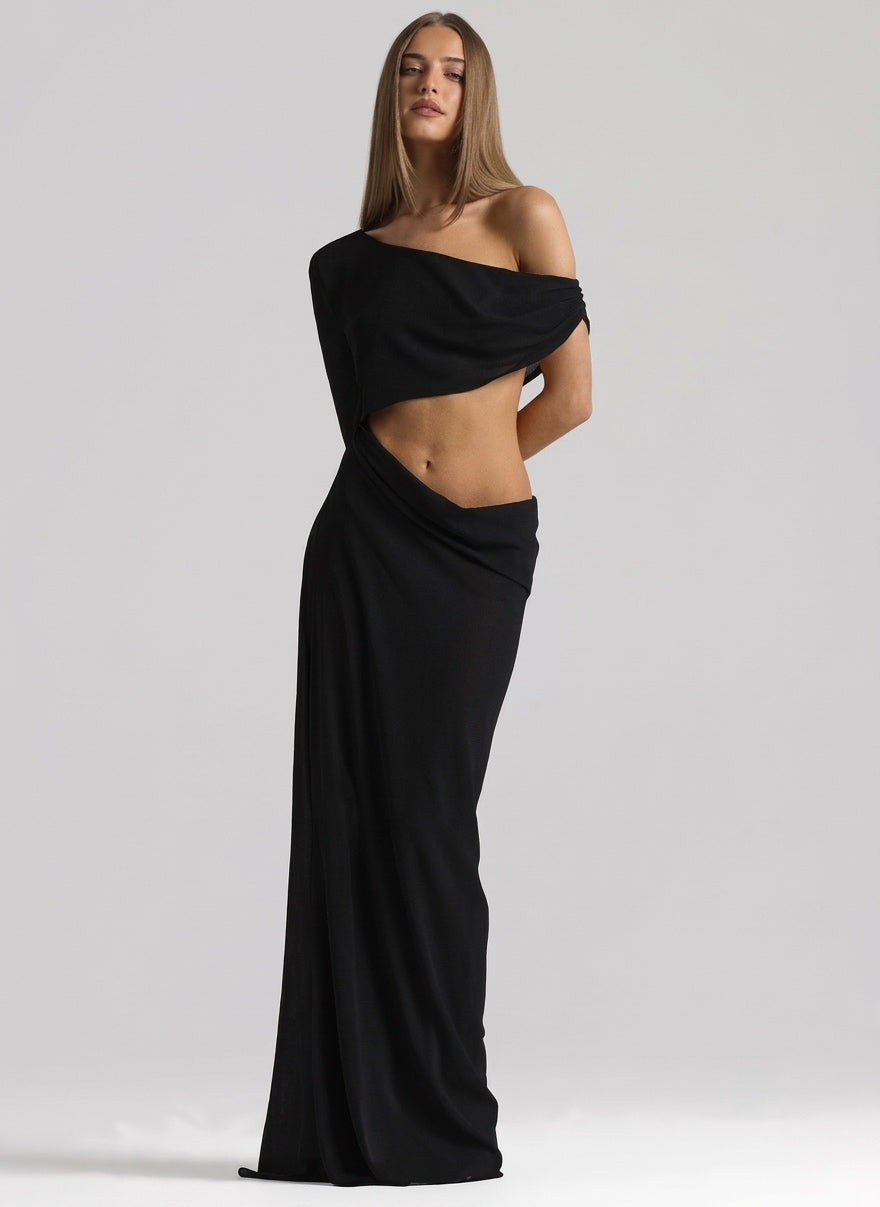 A woman modeling a floor-length black off-the-shoulder gown with a draped cutout at the waist, photographed against a light gray studio background.