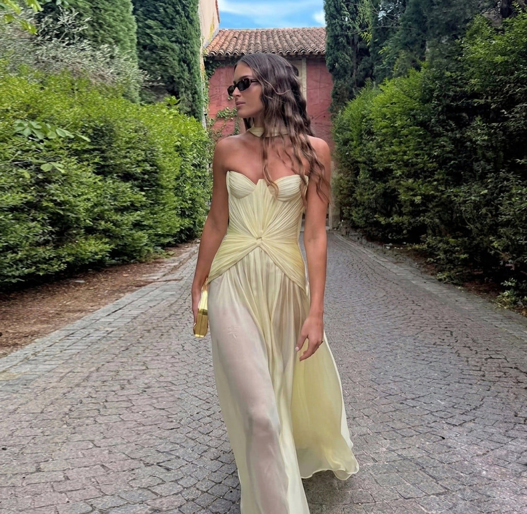 Woman in a light yellow dress standing on a cobblestone path with greenery around.