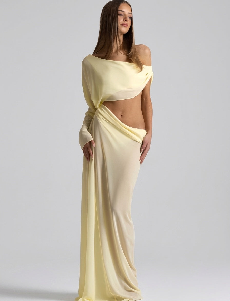 A woman modeling a pale yellow off-the-shoulder maxi dress with a draped wrap-style skirt and a matching draped crop top, photographed against a light gray studio background.
