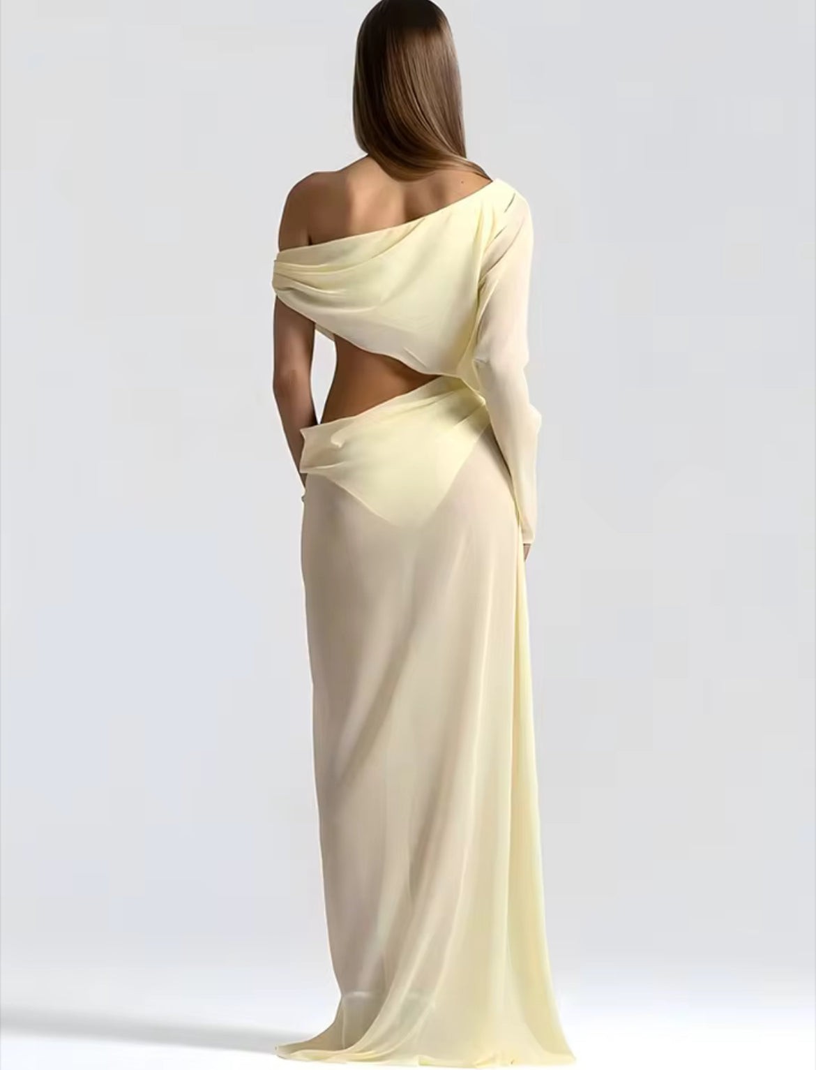 A woman shown from the back wearing a flowing one-shoulder maxi dress in a pale pastel yellow, set against a plain light gray studio background.