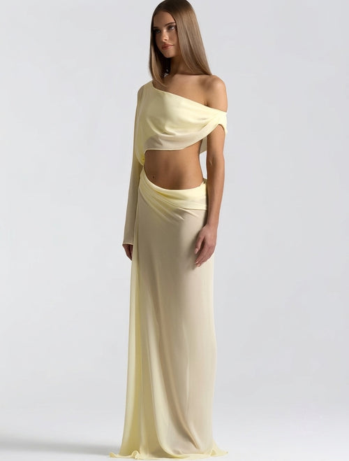 A woman modeling a pale yellow off-the-shoulder maxi dress with a draped cutout waist and long flowing skirt, photographed against a light gray studio background.
