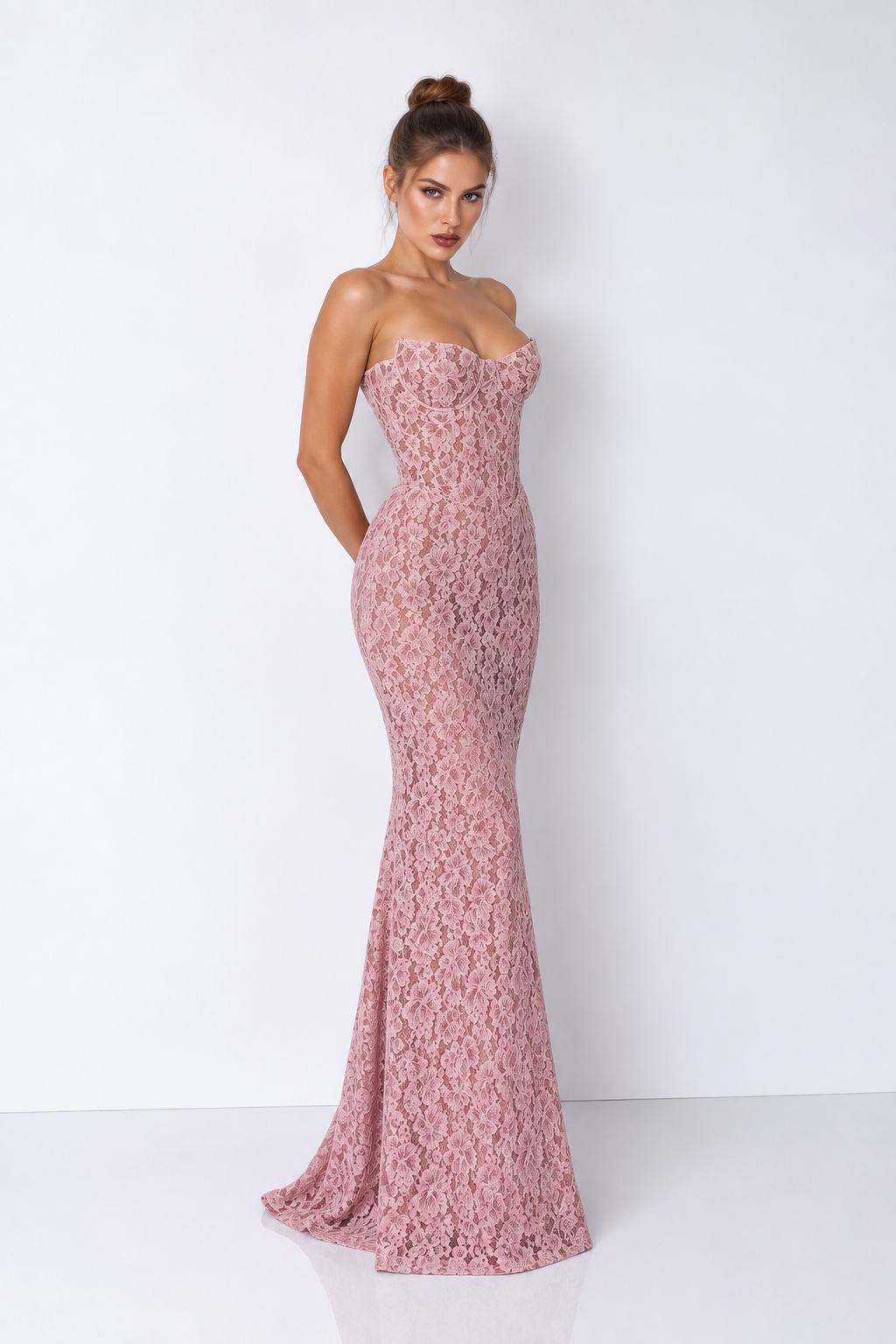 A woman modeling a strapless, floor-length mermaid gown in a dusty rose pink lace fabric against a plain white studio background.