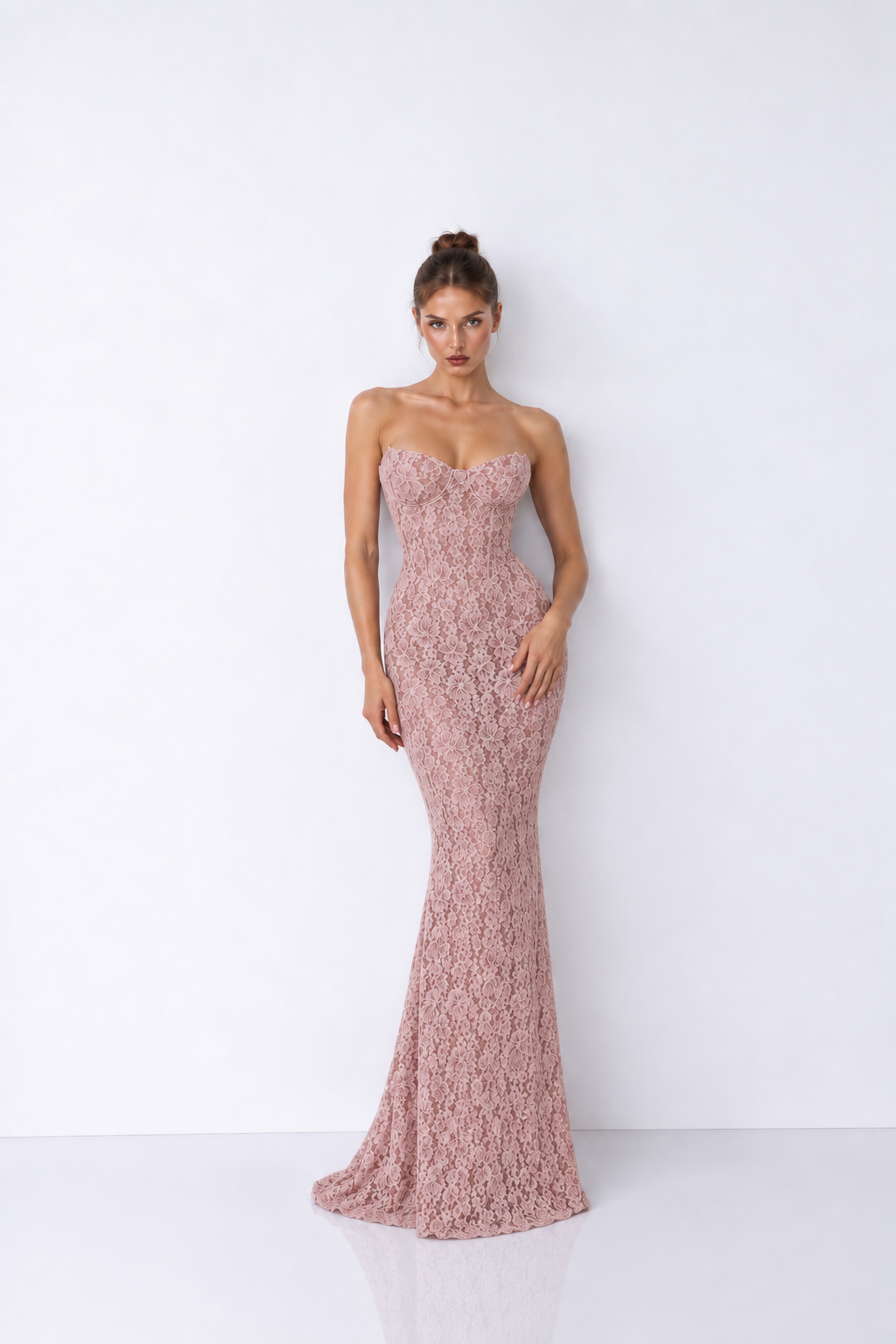 A woman modeling a strapless, floor-length lace gown in a dusty rose pink color, standing against a plain white studio background.