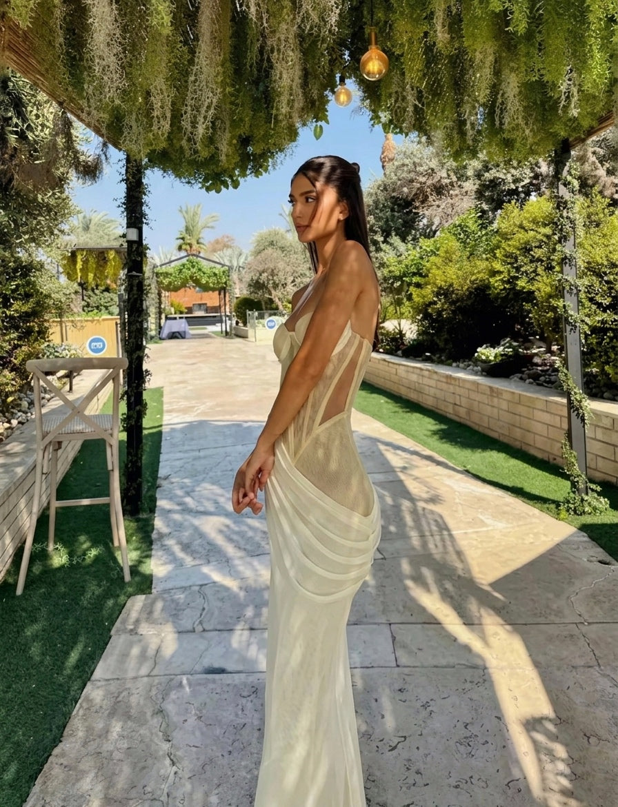 Woman in a white dress standing outdoors with greenery and a clear sky.